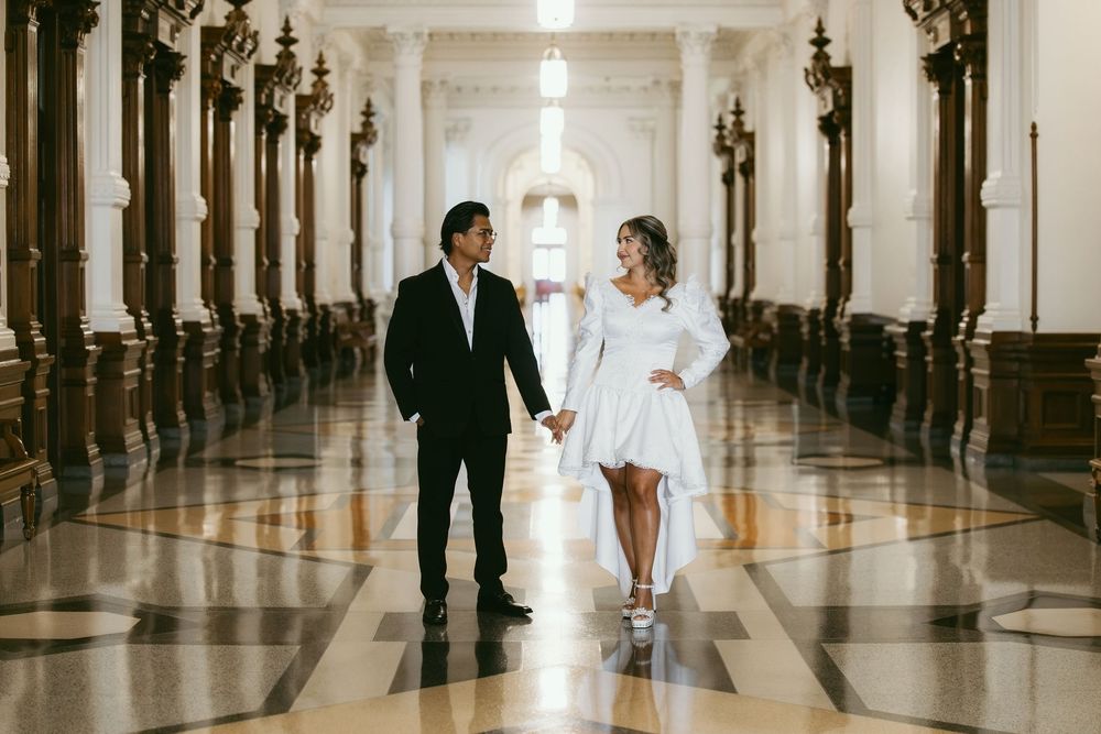 Couple holding hands in elegant marble hallway with ornate columns, woman in white dress, man in black suit.
