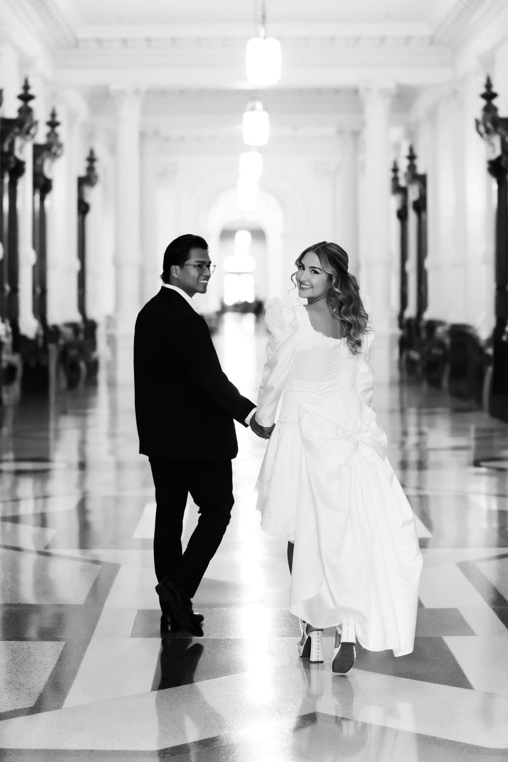 Black and white photo of a bride and groom holding hands walking through an elegant columned hall.
