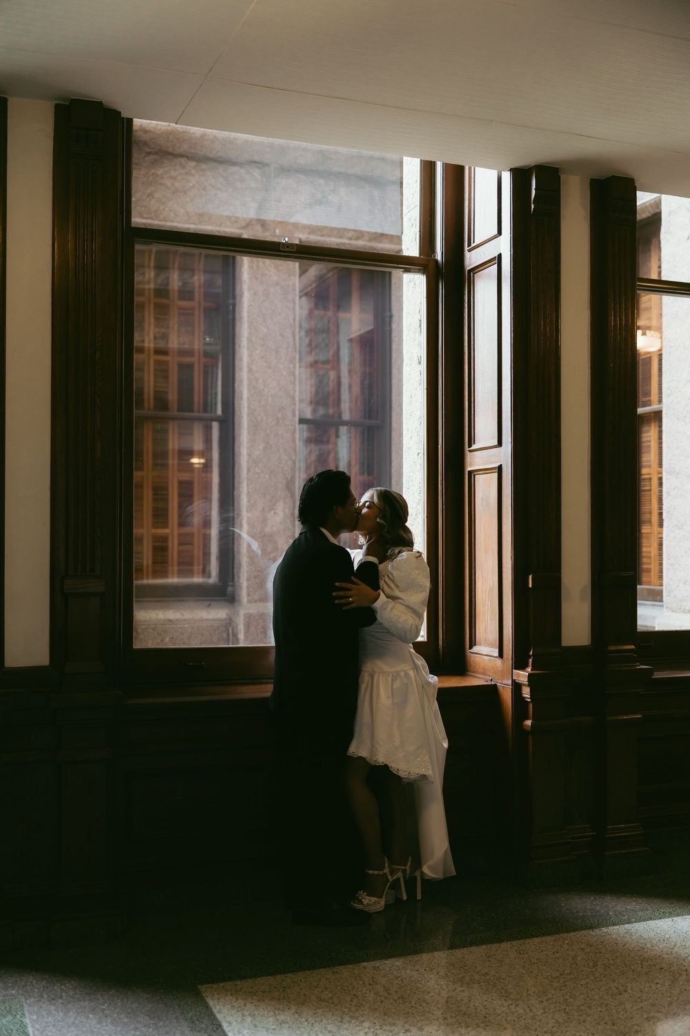 Couple sharing a romantic kiss by large windows in an elegant indoor venue with dark wood trim.
