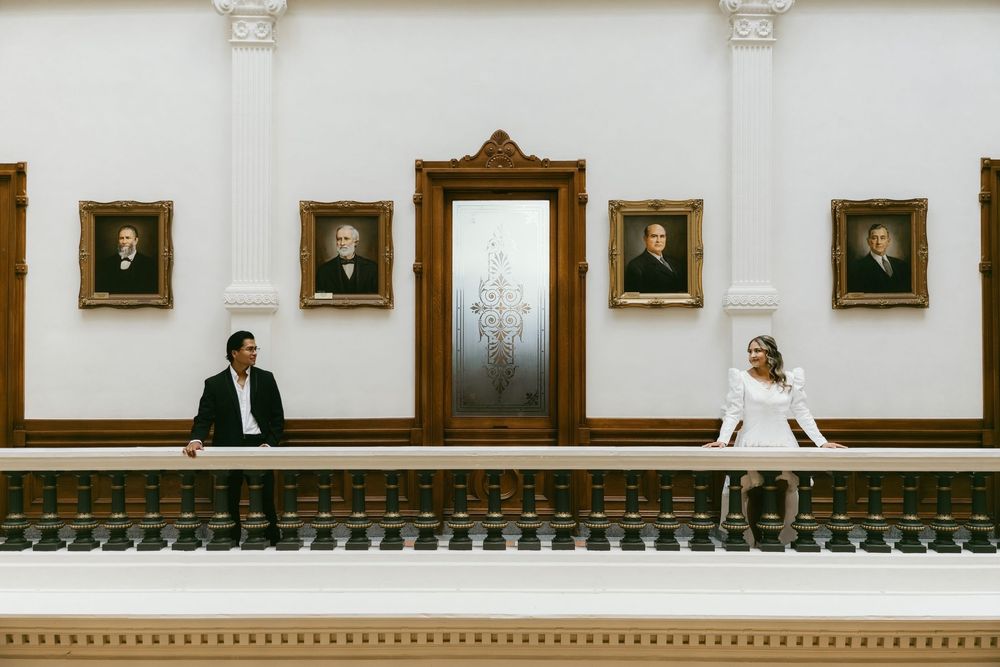 Couple in formal attire sitting apart in elegant hall with portrait paintings on white walls.