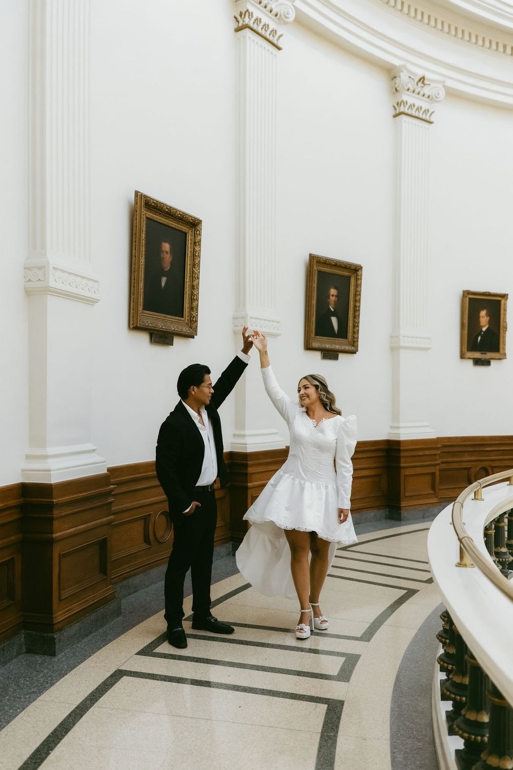 Couple dancing in elegant white marble hall with portrait paintings, bride in short white dress, groom in black suit.