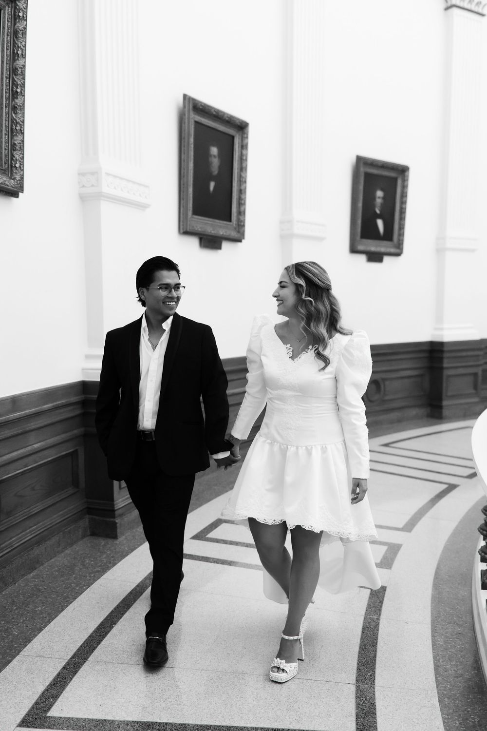 Black and white photo of a couple in wedding attire walking and smiling in an elegant hall with portrait paintings.
