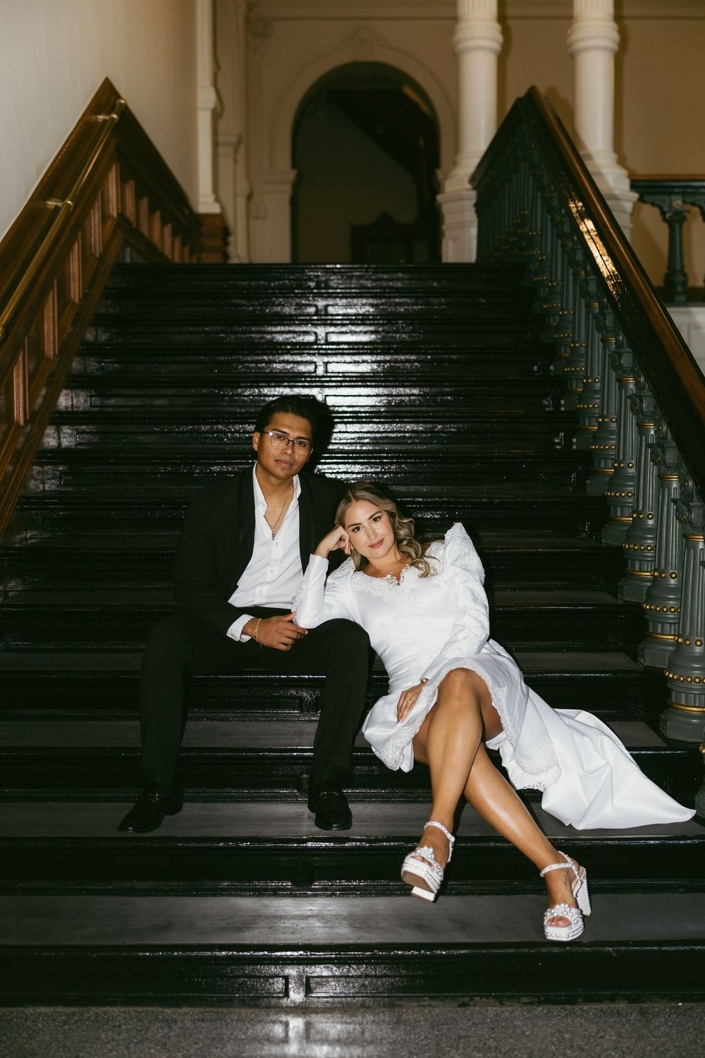 Bride and groom sitting together on grand black staircase in a historic building after wedding.