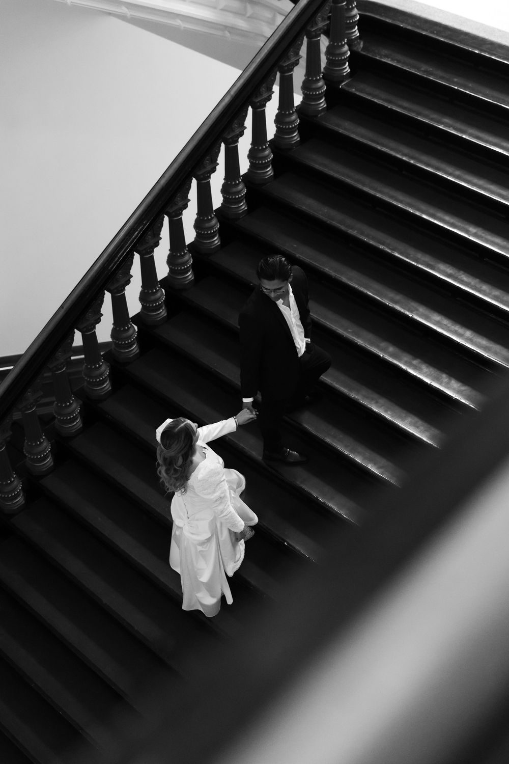Black and white aerial view of bride and groom holding hands on elegant staircase with ornate balusters.