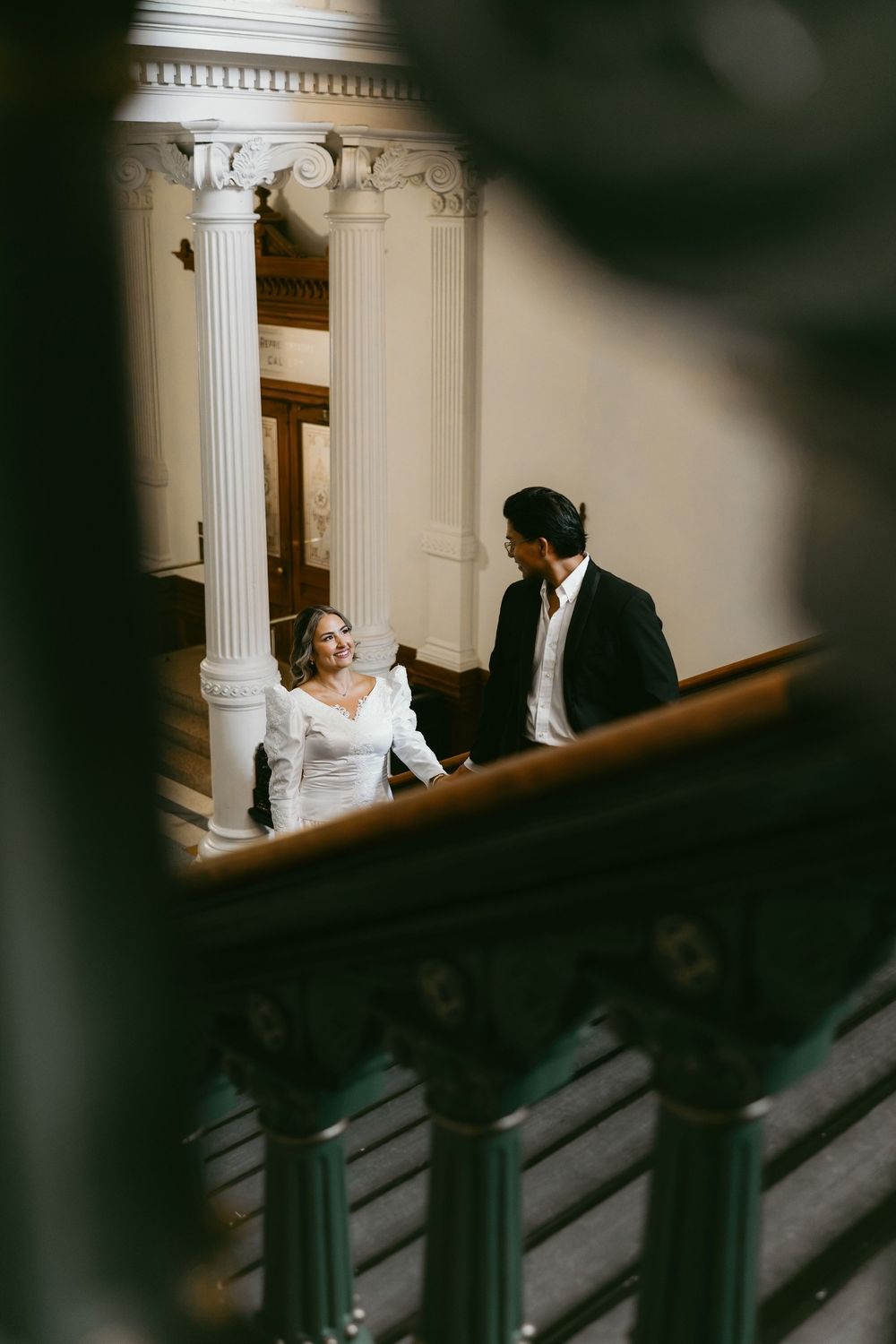 Bride and groom share a romantic moment on grand staircase with white columns in elegant wedding venue.