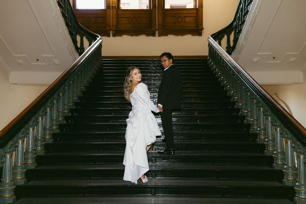 Bride and groom holding hands on elegant dark staircase, bride in white gown looking back, groom in black suit.