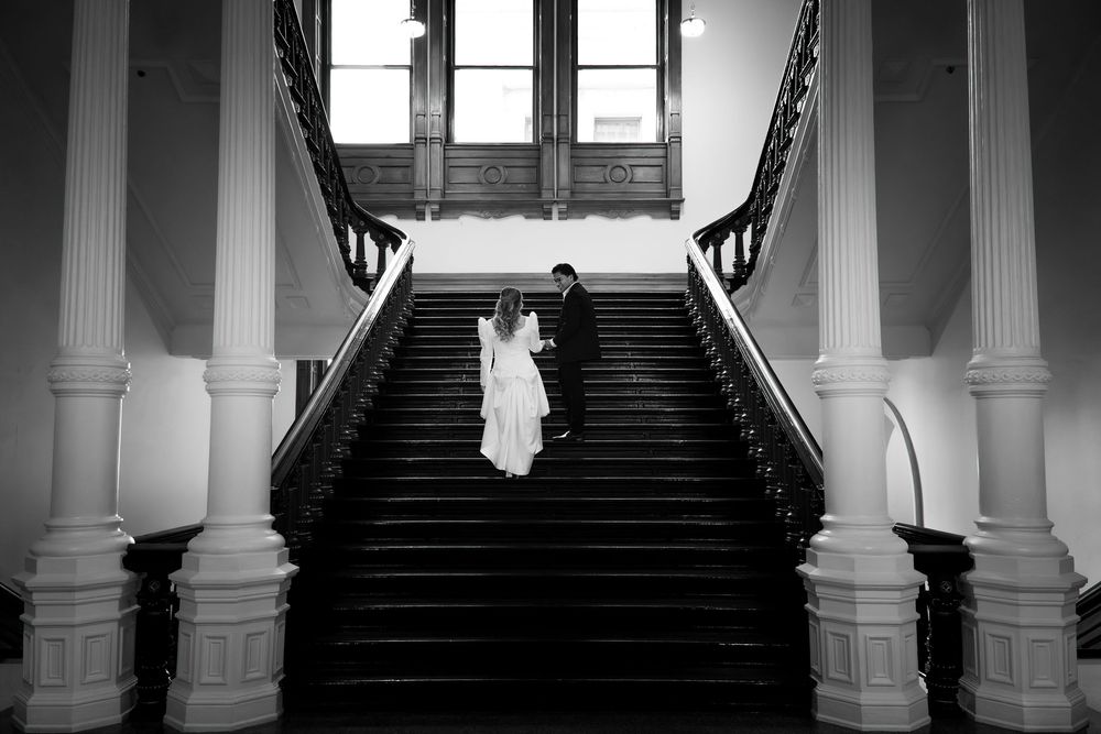 Black and white photo of a bride and groom holding hands on a grand staircase during their wedding day.
