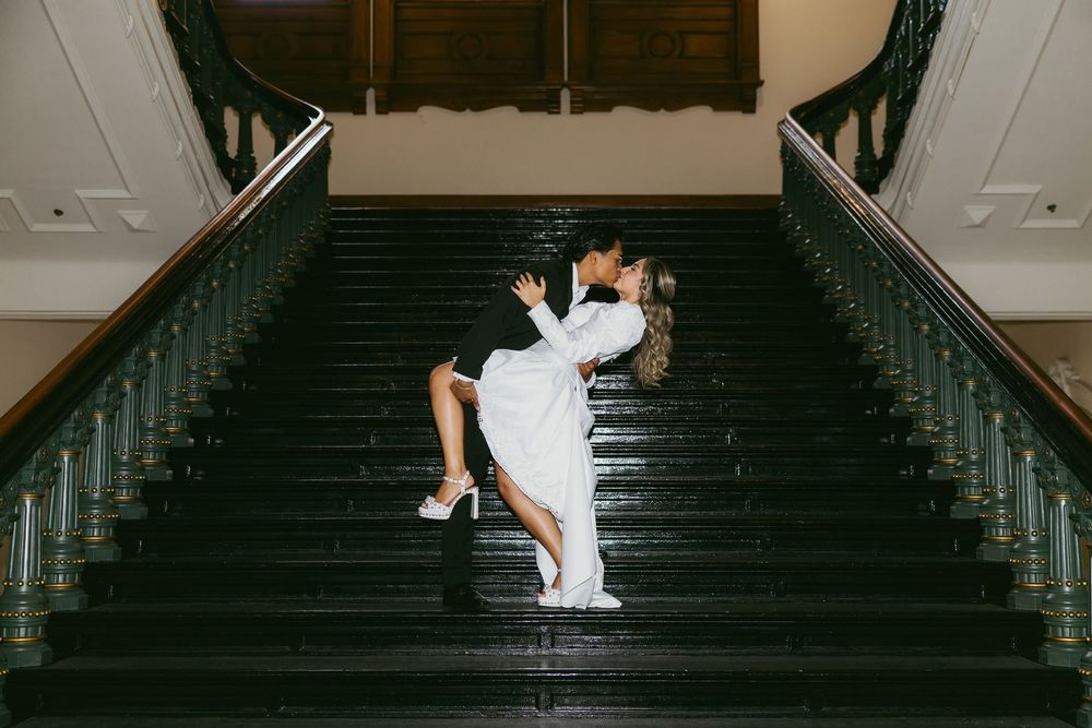Couple sharing a romantic dip kiss on elegant dark staircase in wedding attire.