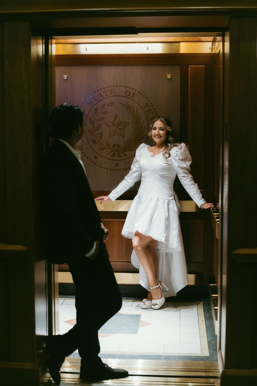 Bride in white dress poses in Texas State Capitol elevator doorway as groom approaches.