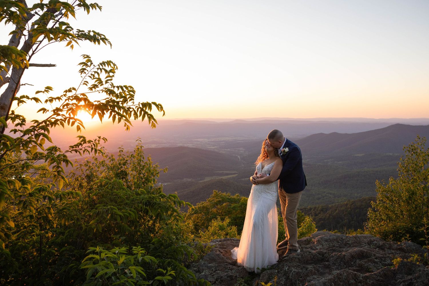 Couple embracing at sunset on a mountain overlook, bride in white gown, groom in navy suit, scenic valley views.