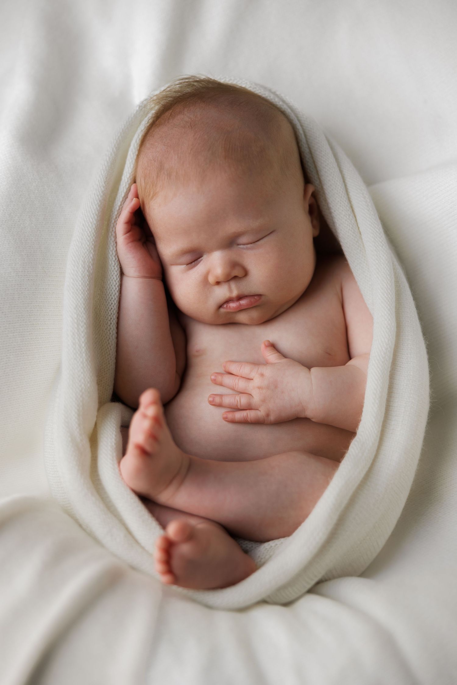 Peaceful newborn baby wrapped in soft white blanket, sleeping curled up in fetal position on white background.
