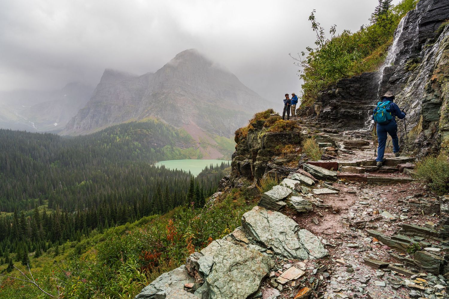 Backpacker with hiking up rocky trail along a bluff line towards two people enjoying the view. Onlookers are gazing at a glacier lake below
