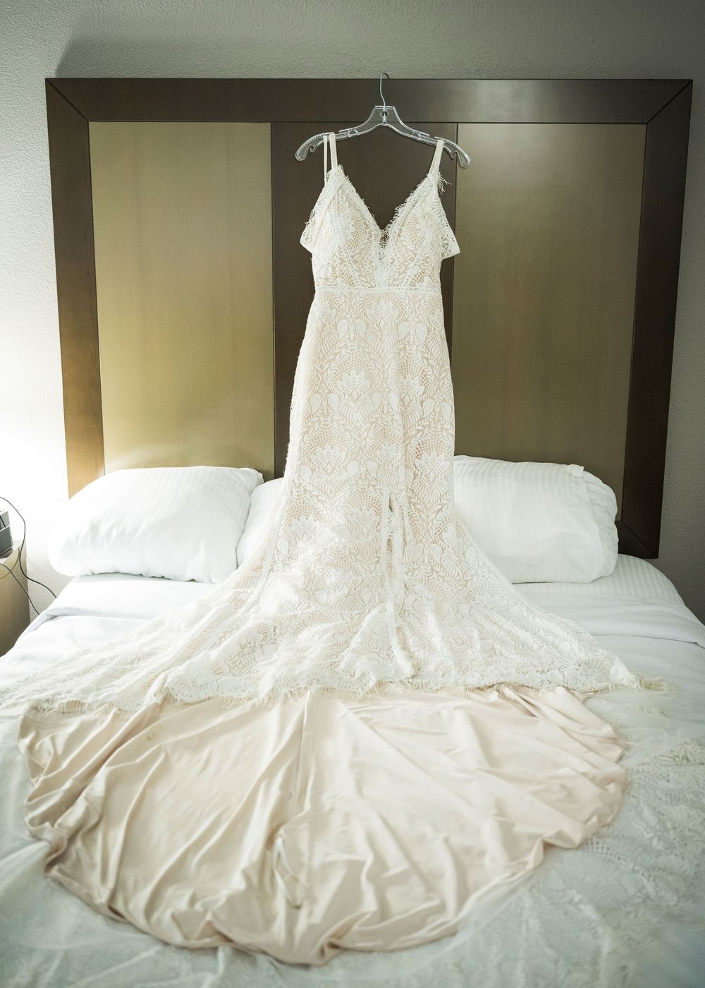 Elegant lace wedding gown with a long train hanging on a hanger over a hotel bed before the ceremony.