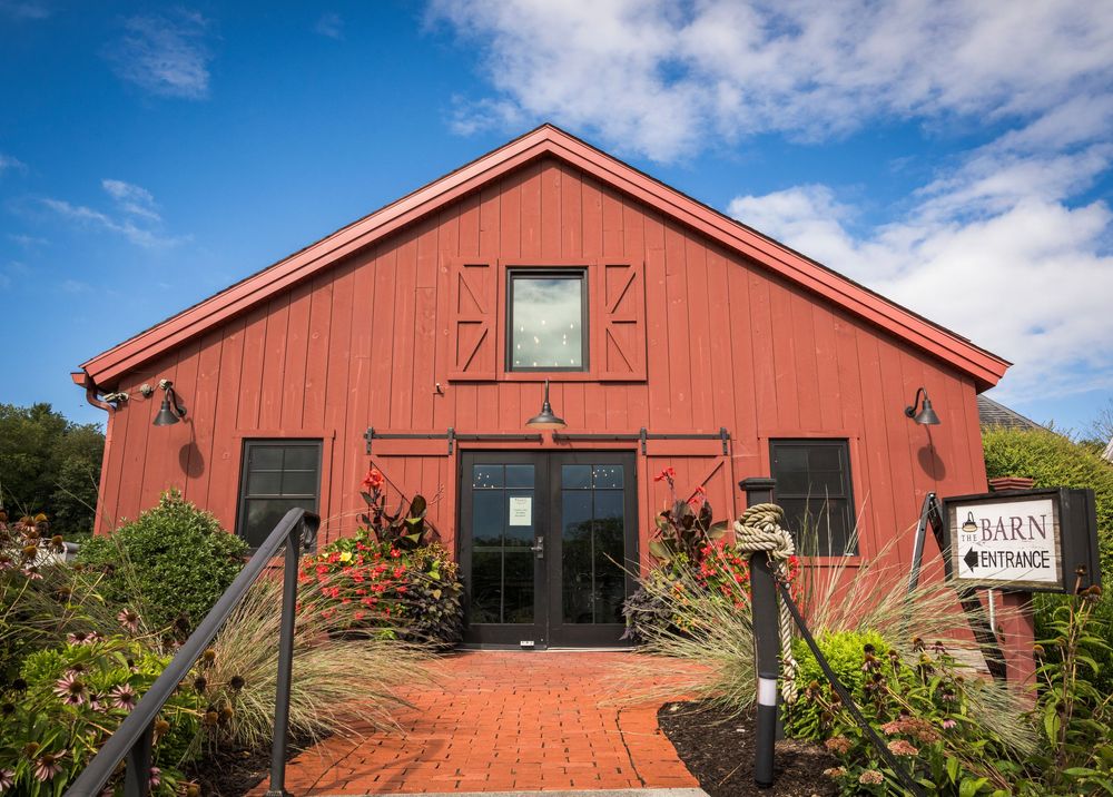 Red barn wedding venue with brick pathway entrance surrounded by lush greenery and flowers.