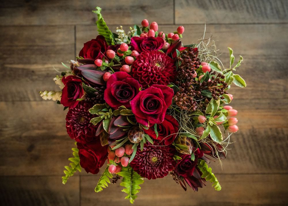 Overhead view of lush red wedding bouquet with roses, dahlias, and berries on wood surface.
