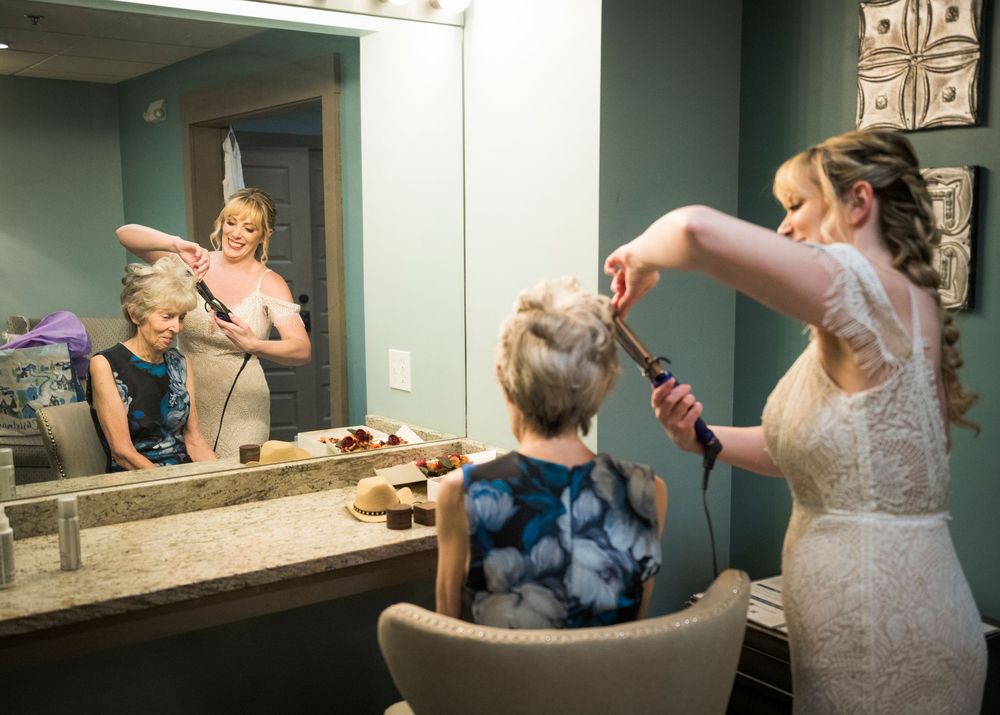 Bride getting hair styled with curling iron while bridesmaids assist in mirror reflection.