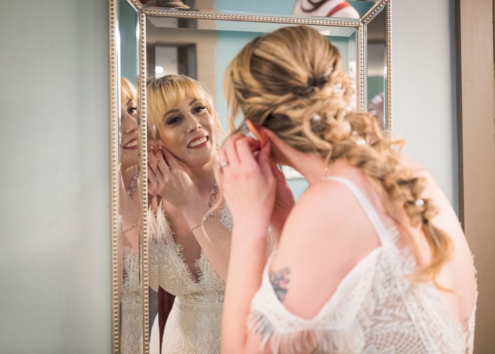 Bride in white lace gown adjusting earring while smiling at her reflection in an ornate gold mirror.