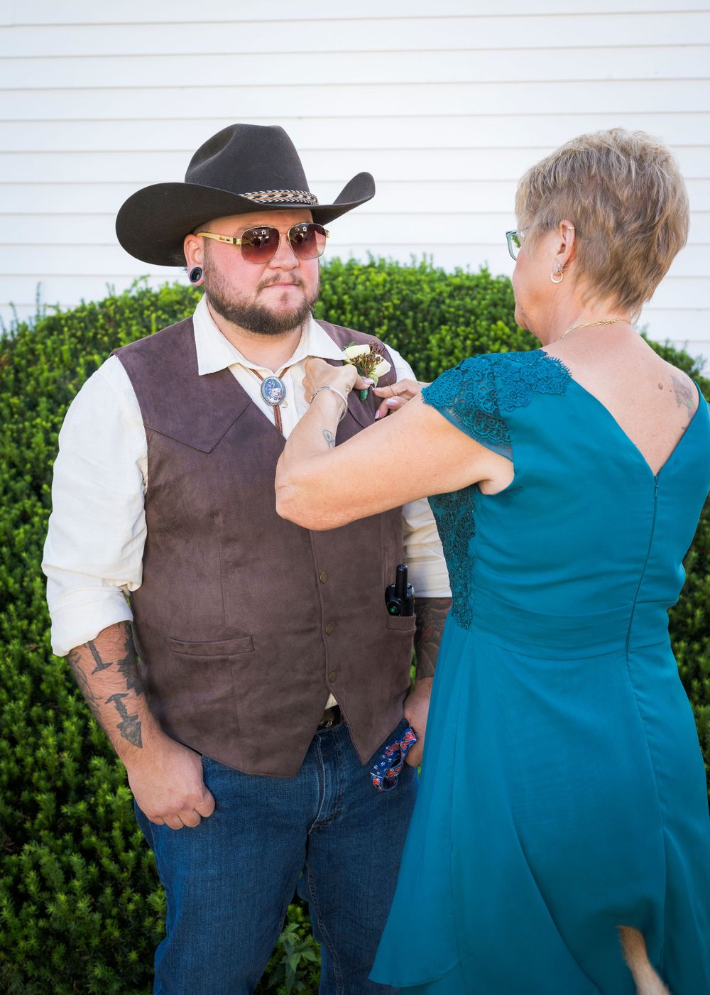 Woman in teal dress pins boutonniere on groom wearing brown vest and black cowboy hat.