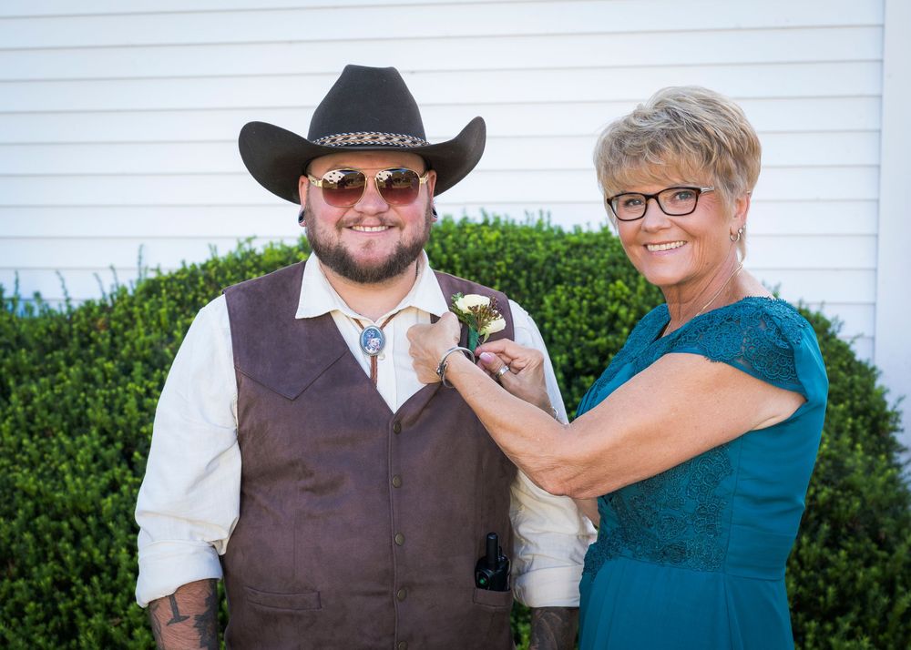 Woman in teal dress pins boutonniere on man in cowboy hat and brown vest at outdoor wedding.