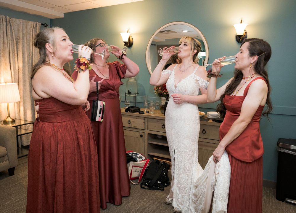 Bride in white gown toasts champagne with three bridesmaids in burgundy dresses before wedding.