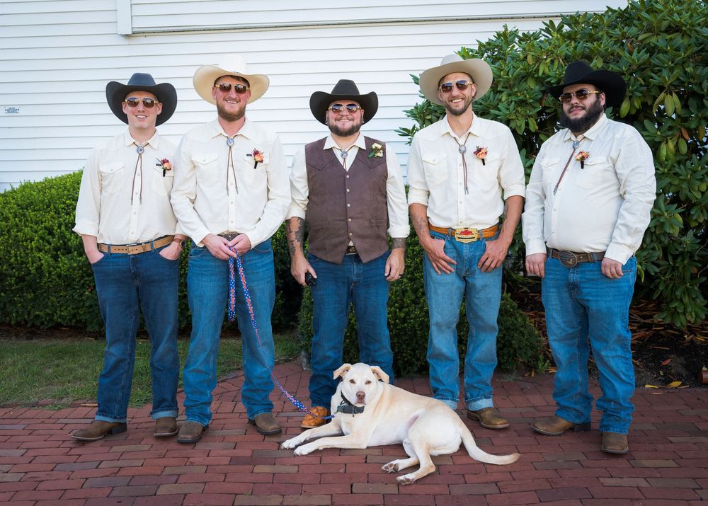 Five groomsmen in cowboy hats and jeans posing with a yellow Labrador dog at a rustic country wedding.