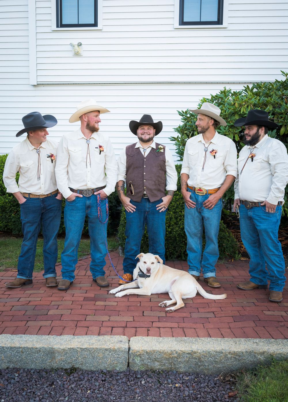 Five groomsmen in cowboy hats, white shirts, and jeans pose with white dog at country wedding.