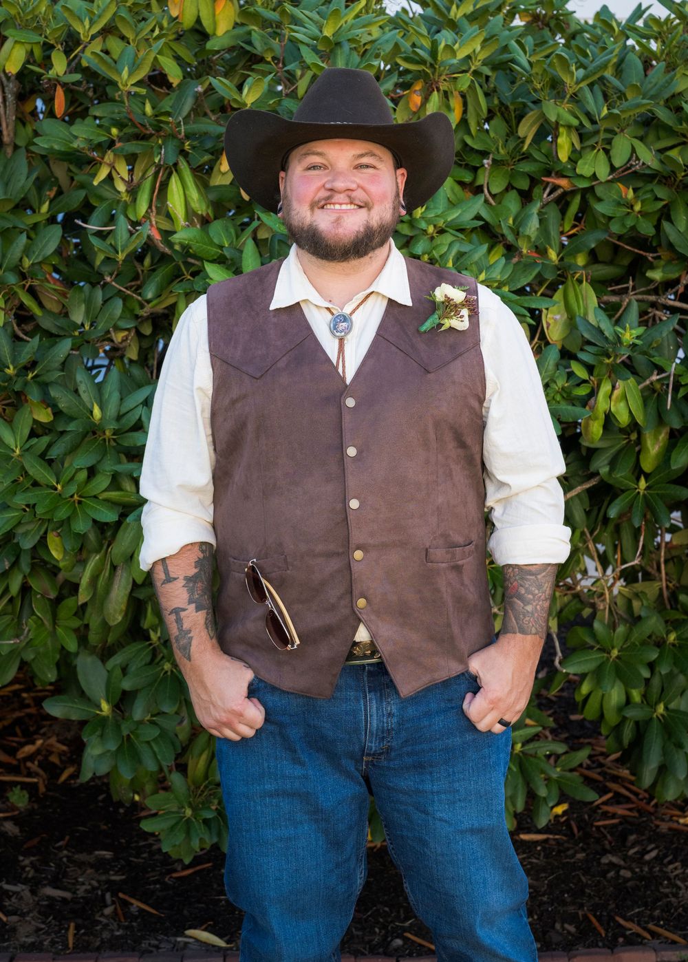 Smiling groom in a brown suede vest, cowboy hat, and boutonniere posing outdoors at his country wedding.