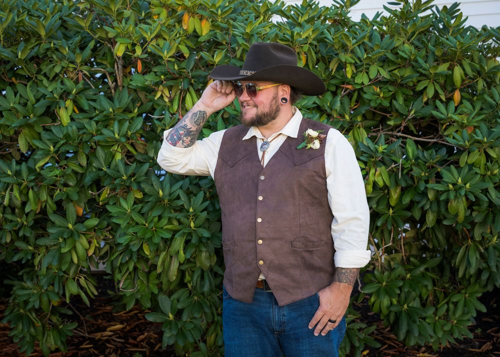 Groom in cowboy hat, brown vest, and boutonniere poses outdoors before rustic wedding ceremony.