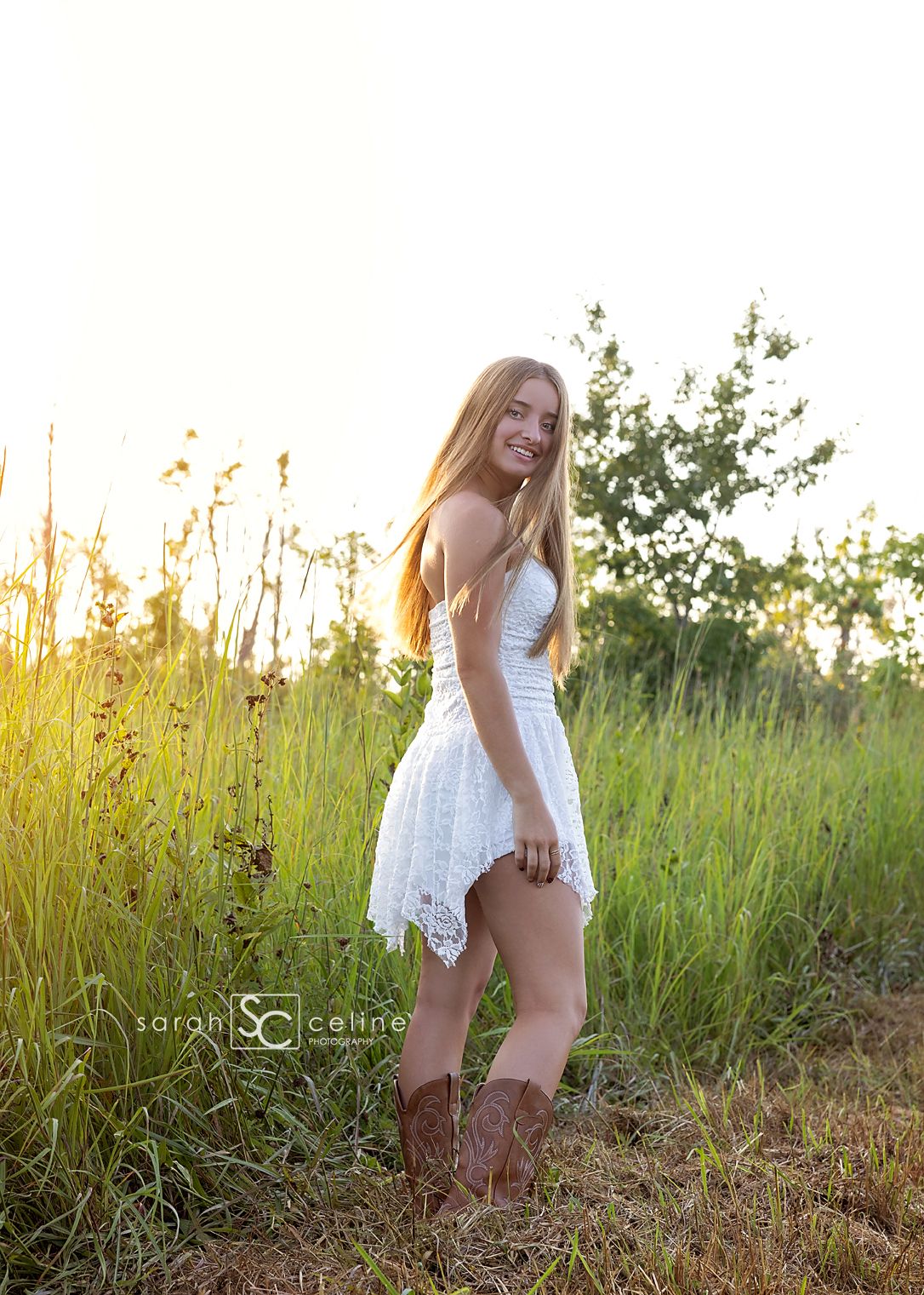 Young woman in white lace dress and cowboy boots posing in sunlit field for senior portrait photo.