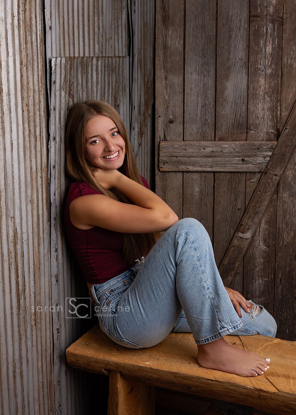 Smiling teenage girl in red top and jeans sitting barefoot against rustic wooden barn wall on a bench.