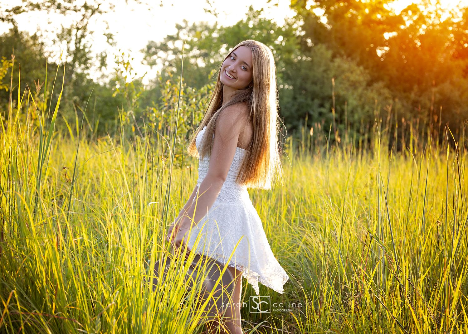 Young blonde woman in white dress standing in golden sunlit meadow with tall green grass and trees in background.