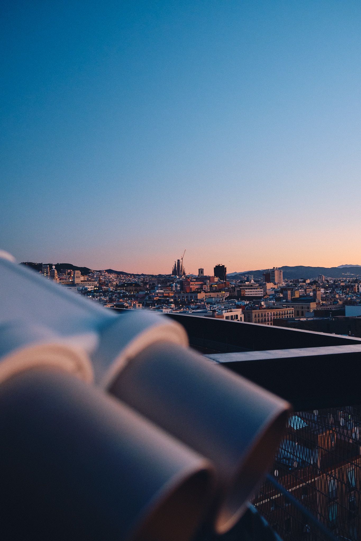 Coin-operated binoculars on a rooftop overlooking Barcelona cityscape at sunset with clear blue sky.