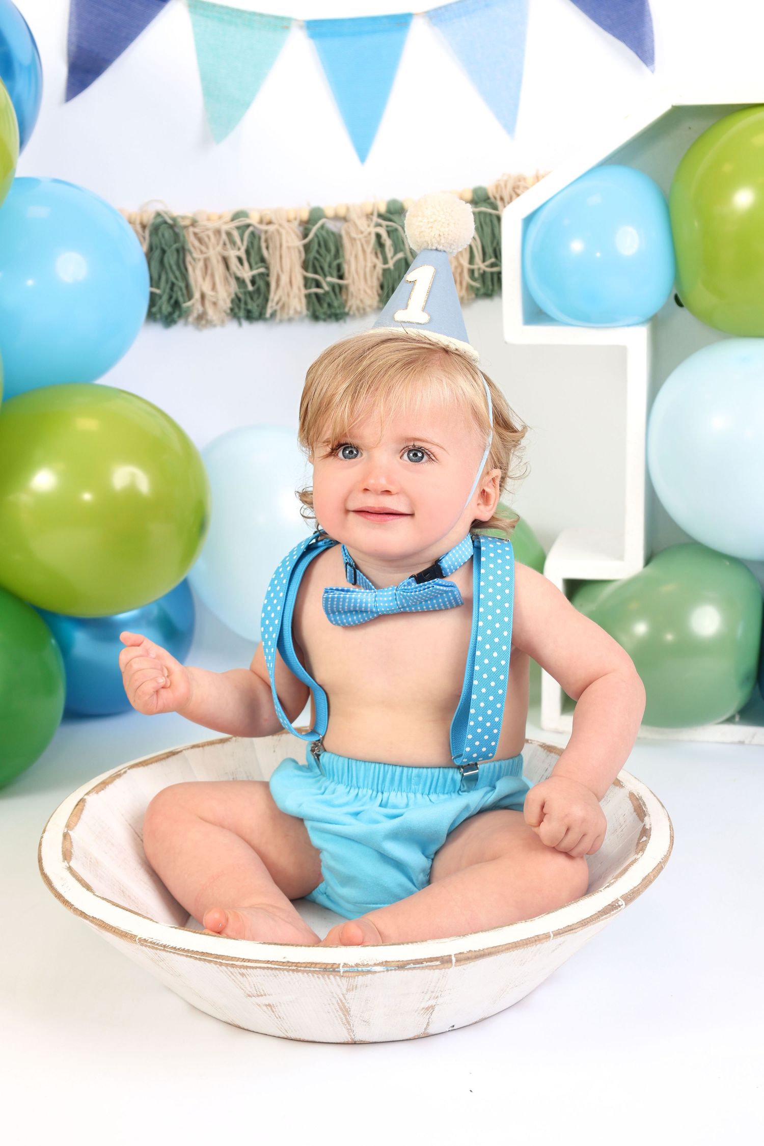 Smiling baby boy in blue suspenders and bow tie sits in a white bowl surrounded by colorful balloons at a first birthday.