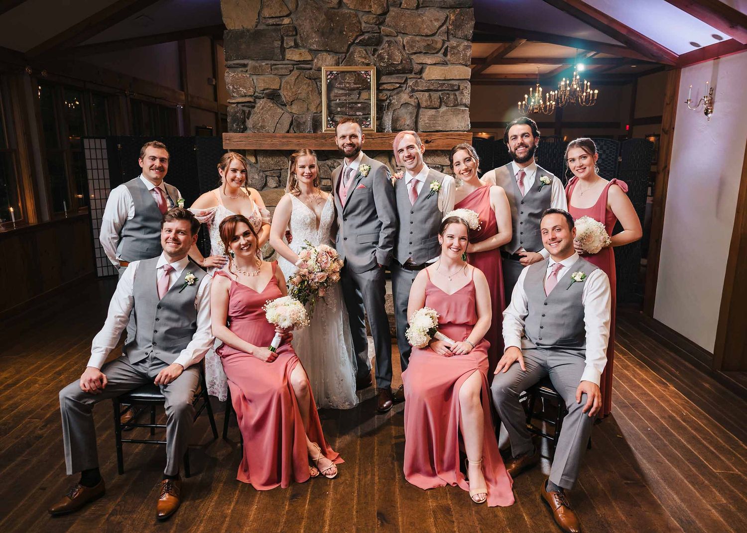 Bride and groom with full bridal party gathered by a stone fireplace at Fernhill Barn in Spencer, Massachusetts.