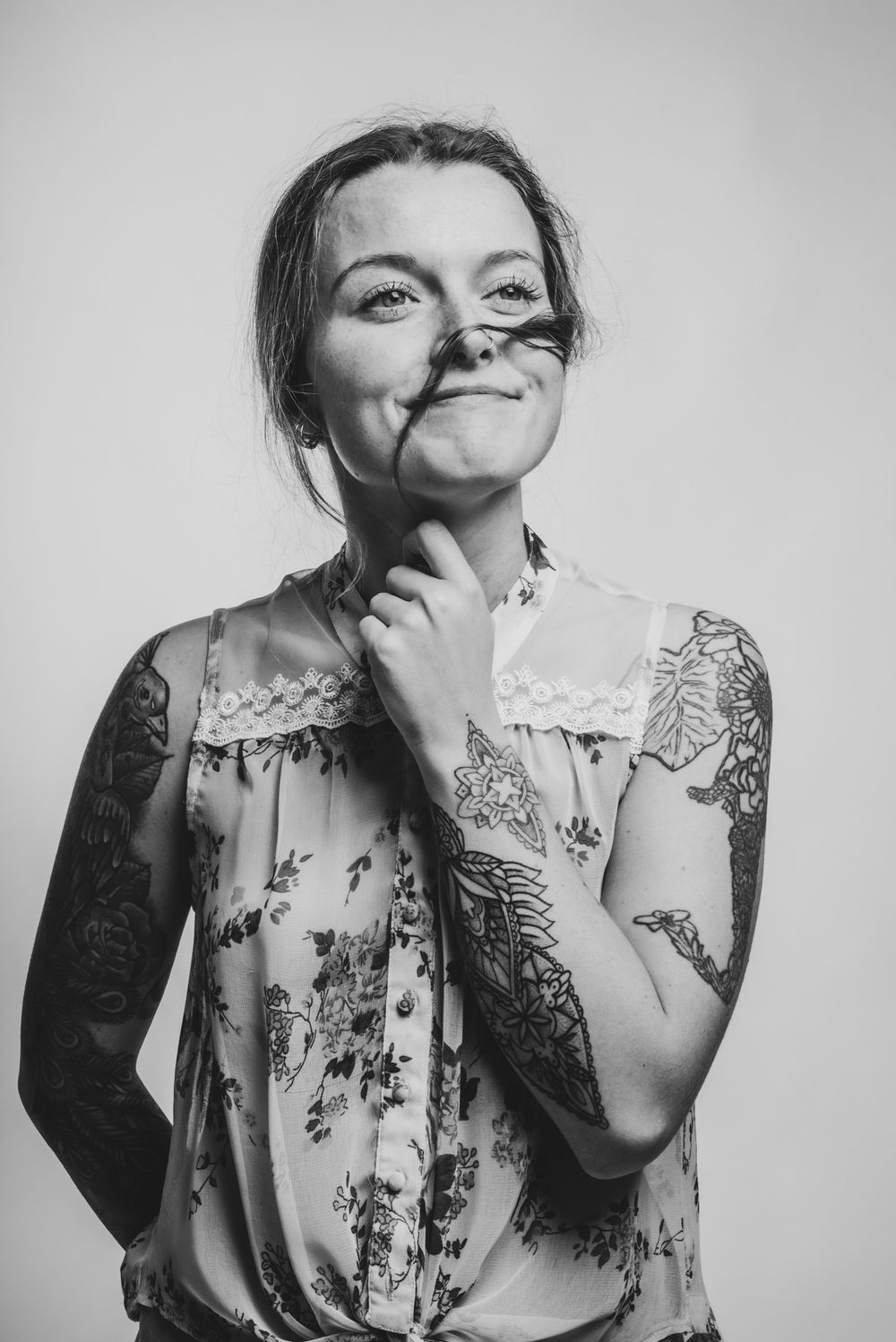 Zwart wit studioportret van een vrouw met een neusring en getatoeëerde armen, met een haarlok speels voor haar gezicht