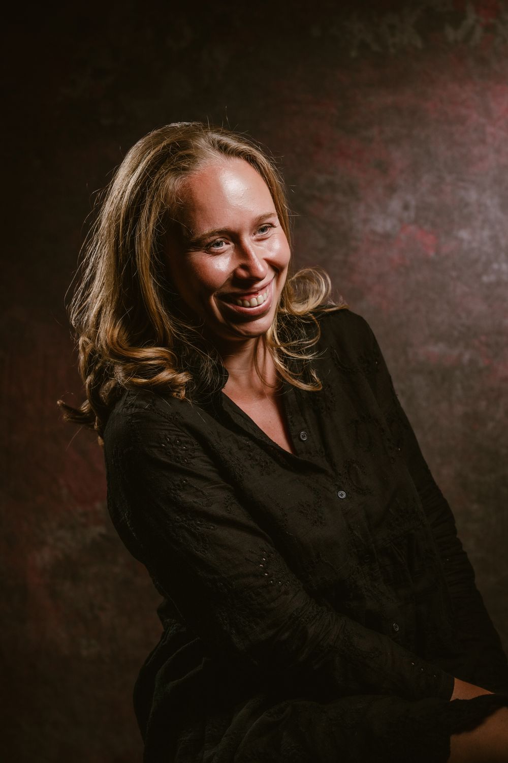 Warm belicht studioportret van een lachende vrouw met lang lichtbruin haar en een zwarte blouse, zittend voor een donkere achtergrond.