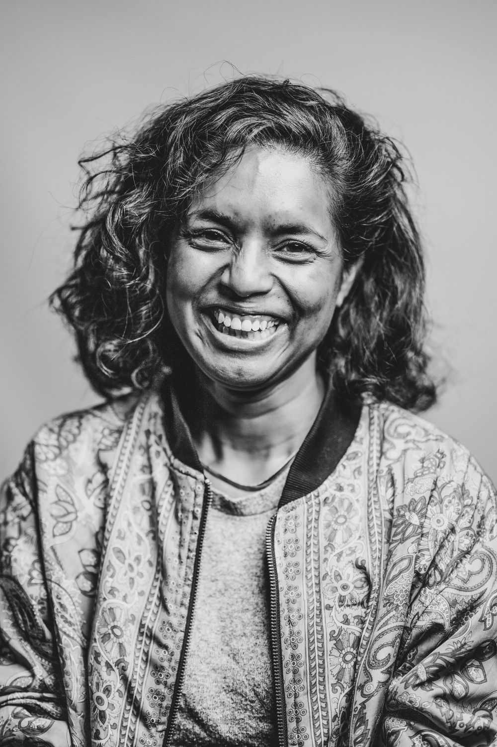 Spontaan zwart wit portret van een breed lachende vrouw met krullend haar en een jas met bloemenmotief bij Bart Boodts