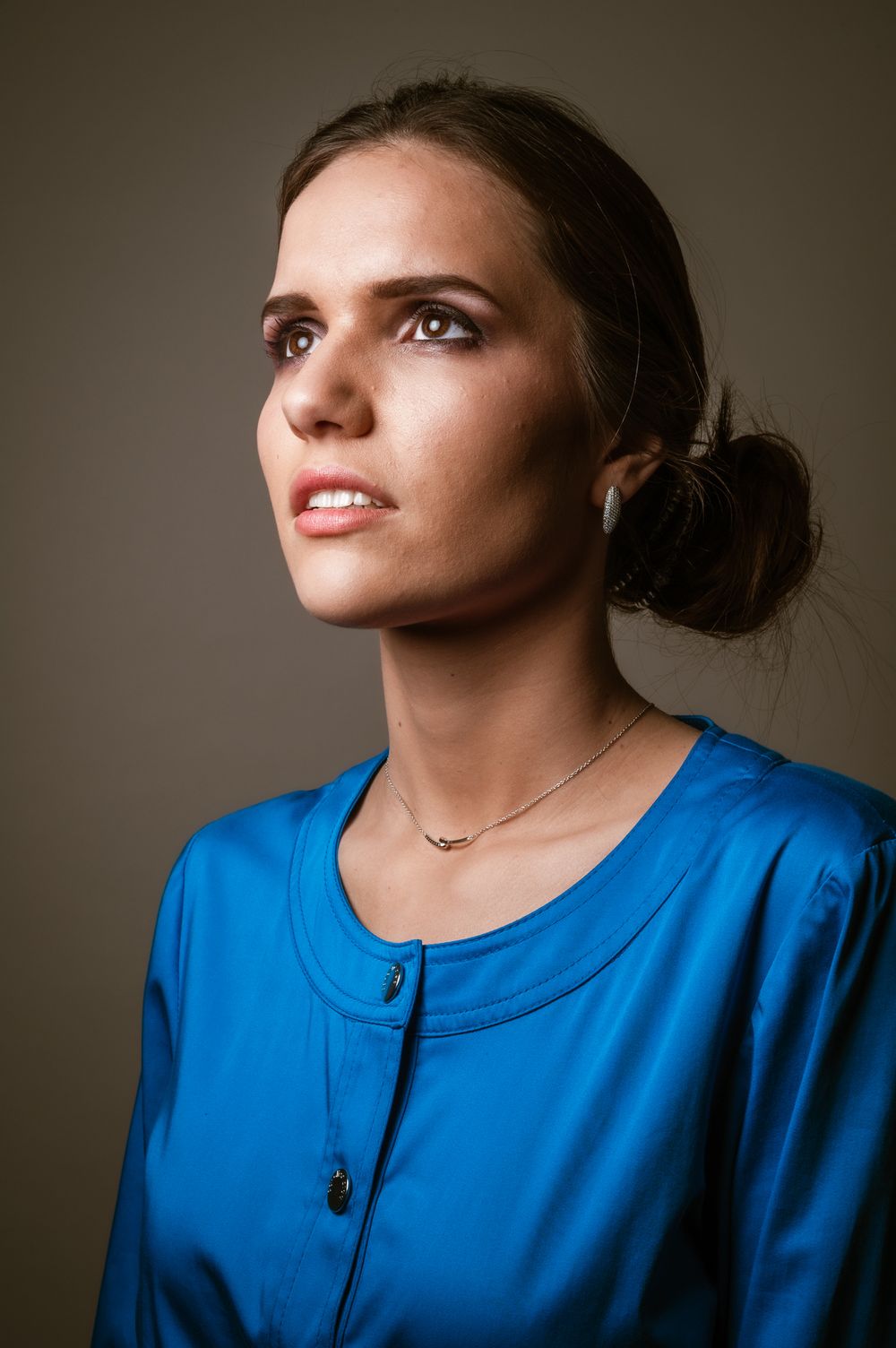 Elegant studioportret van Miss België met opgestoken donker haar en een felblauwe blouse, terwijl ze omhoog kijkt in zacht licht.
