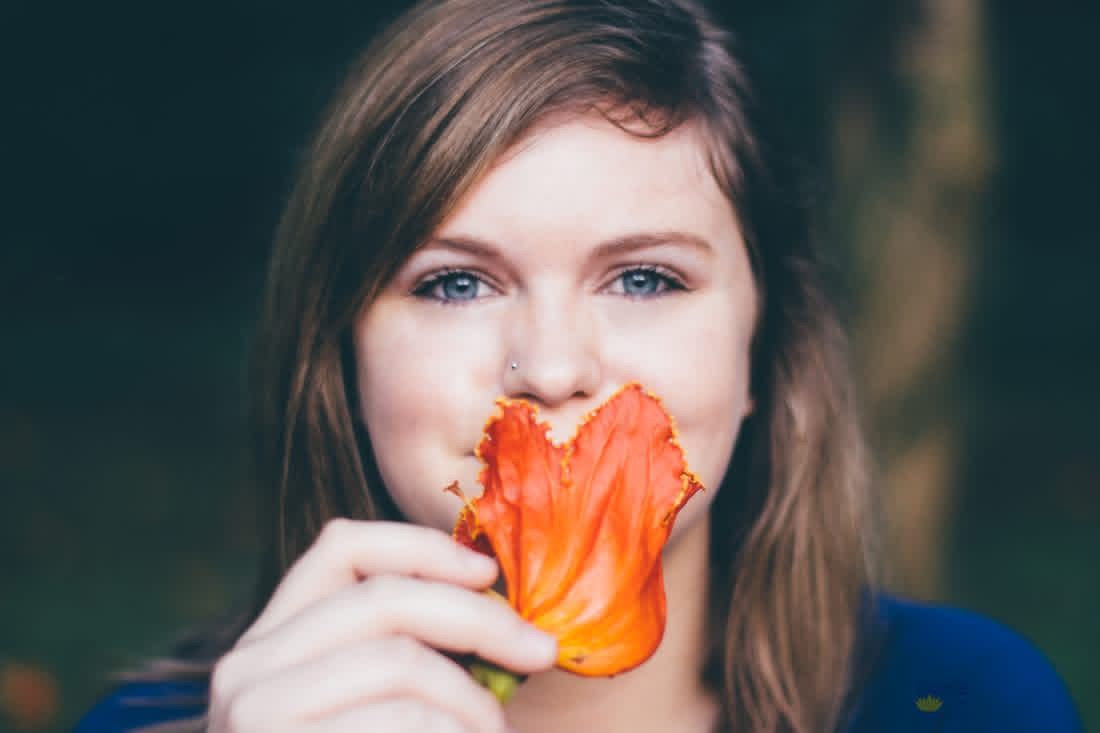 Young woman with blue eyes holding an orange flower up to her face in a soft-focus outdoor setting.