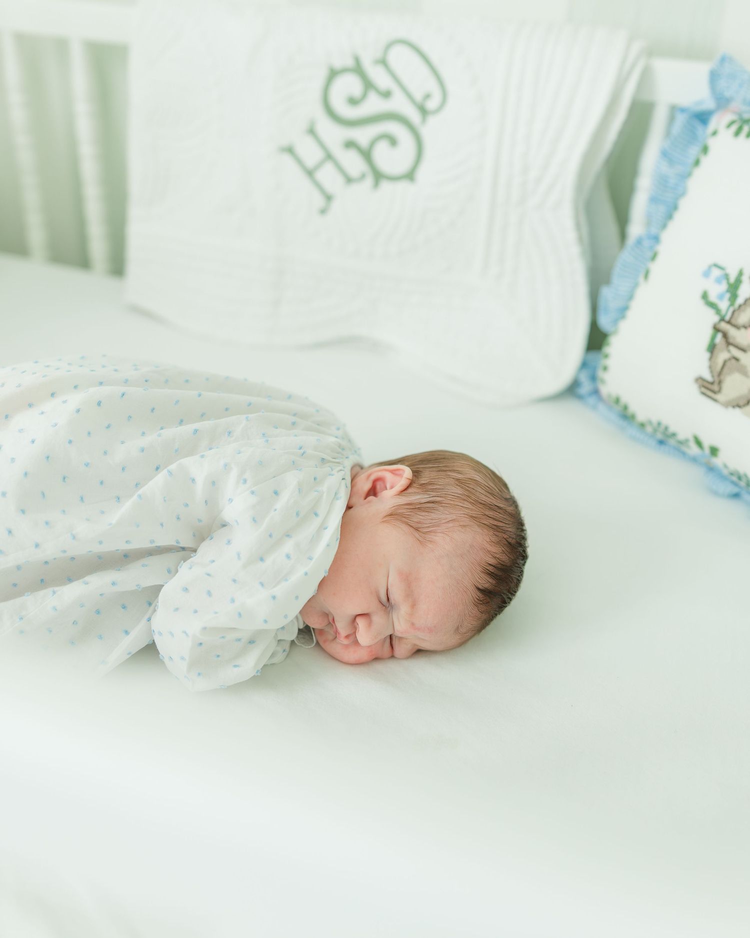 Sleeping newborn baby swaddled in white dotted gown lying in a white crib with monogrammed pillow.