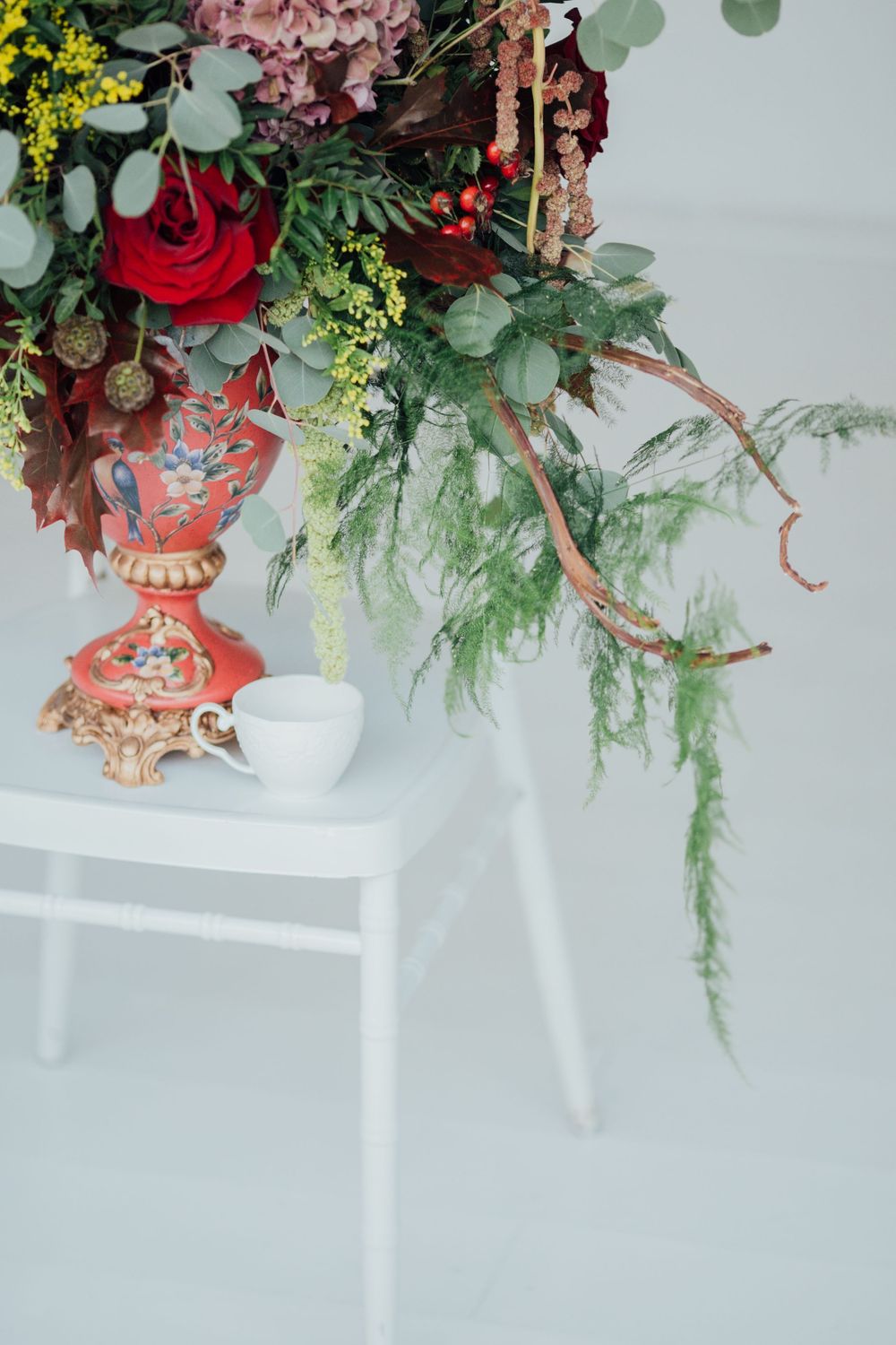 Elegant floral arrangement with red roses and greenery in ornate vase on white chair with teacup.