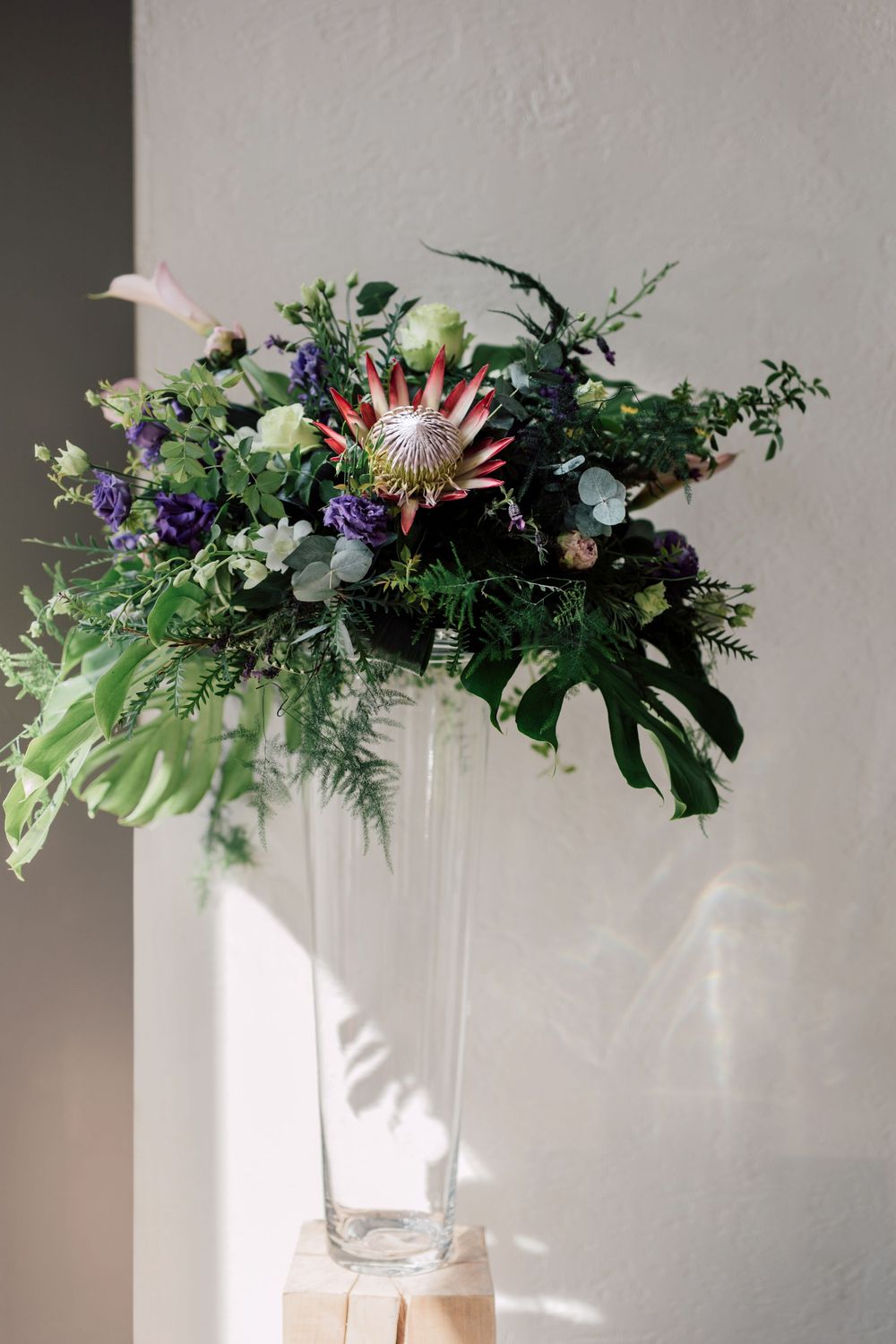 Tall glass vase with lush tropical floral arrangement featuring a pink protea, monstera leaves, and purple blooms.