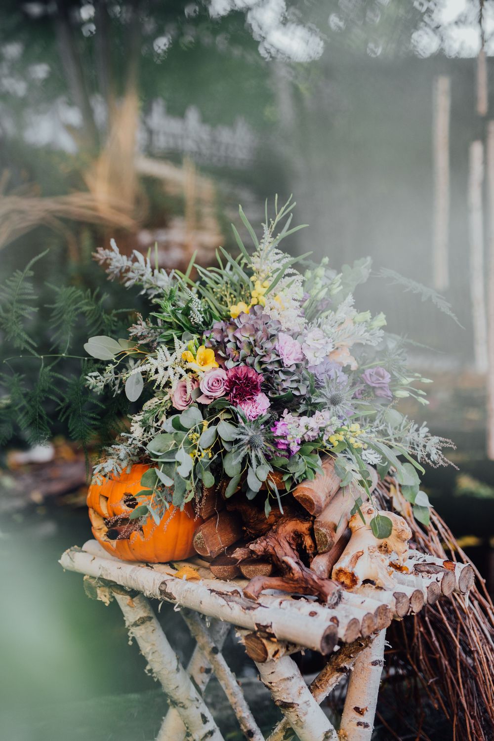 Autumn floral arrangement in a pumpkin vase on a rustic birch wood table with wildflowers and greenery outdoors.