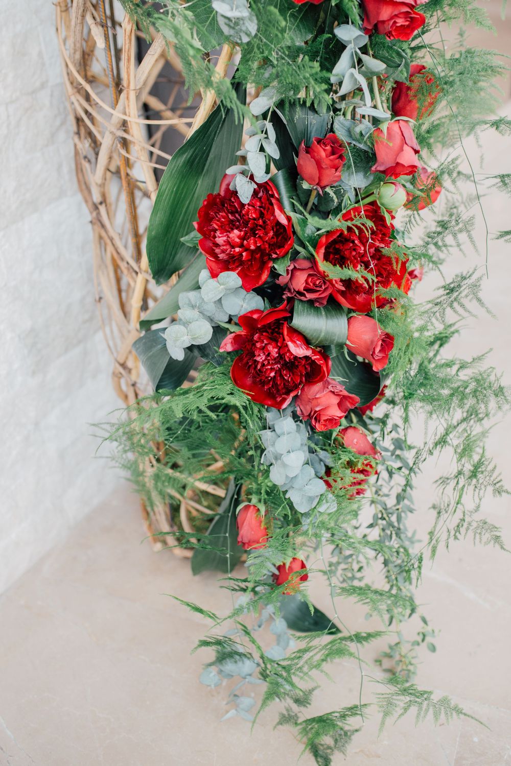 Red peonies and roses with lush greenery arranged on a rustic wicker arch for an elegant wedding decoration.