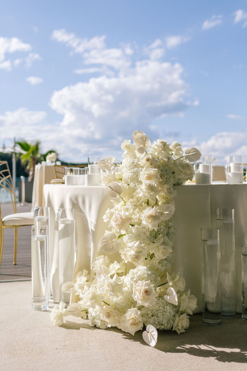 Elegant outdoor wedding reception table adorned with lush white floral arrangement, candles, and white linens under a blue sky.