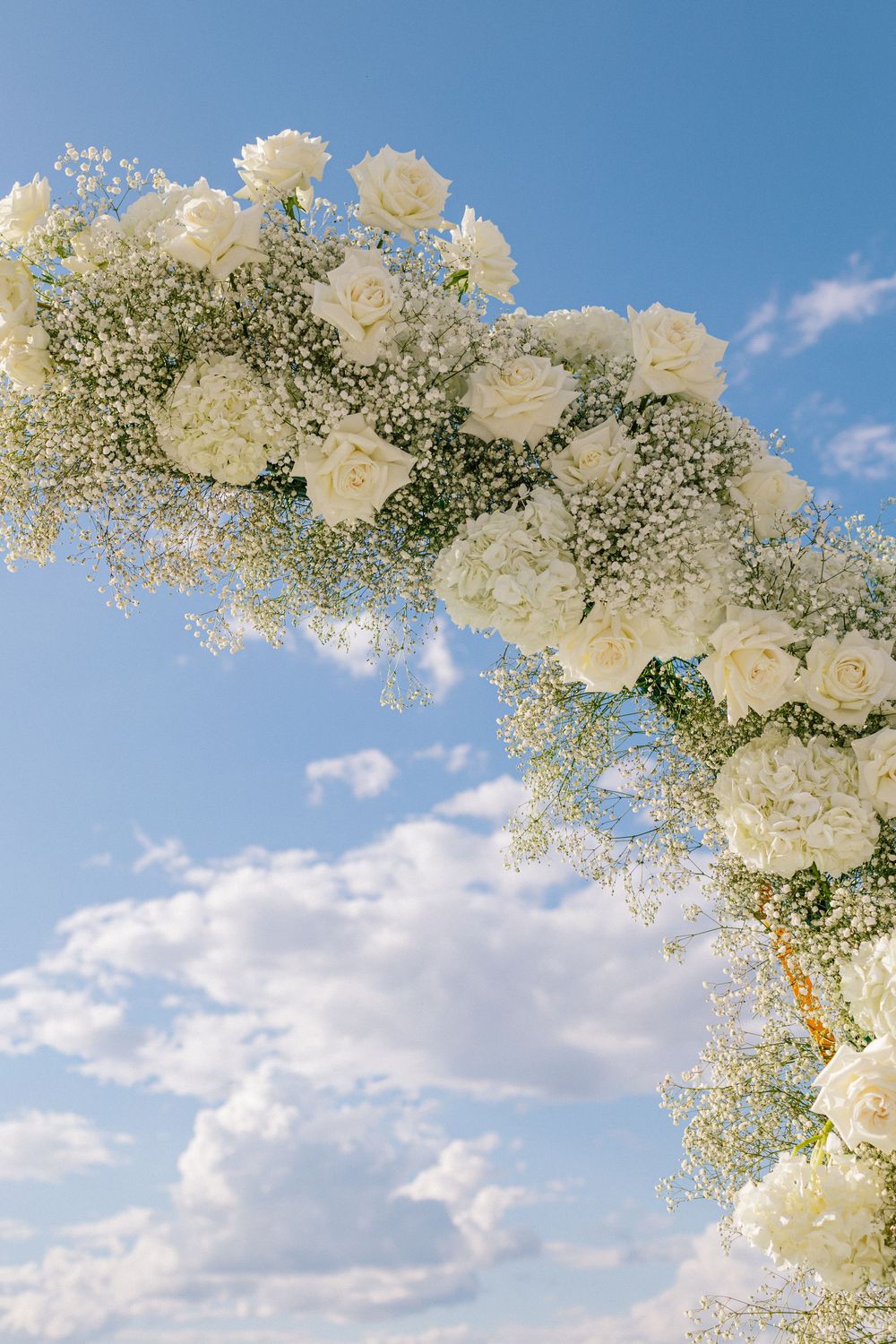 White roses and baby's breath floral arch against a blue sky, perfect for an elegant outdoor wedding ceremony.