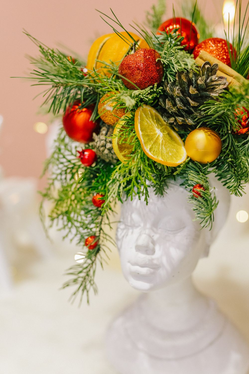 White mannequin head wearing festive Christmas wreath with pine branches, red and gold ornaments, pine cone, and citrus slices.