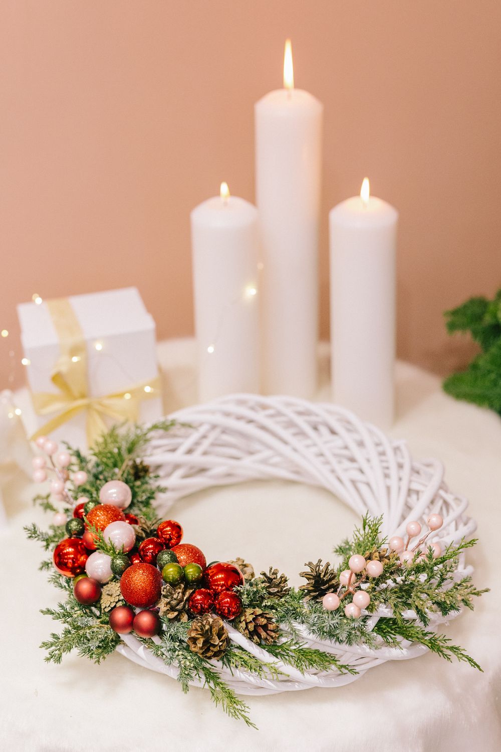 White wicker Christmas wreath with red ornaments, pine cones, and greenery, displayed with lit white candles and a gift box.