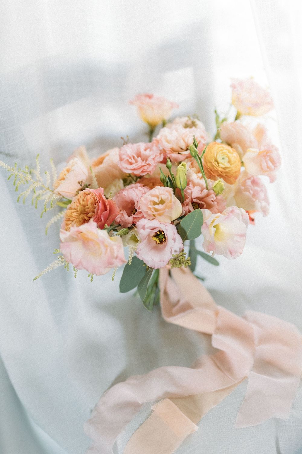 Elegant bridal bouquet of peach, pink, and coral roses with ribbon on soft white fabric.