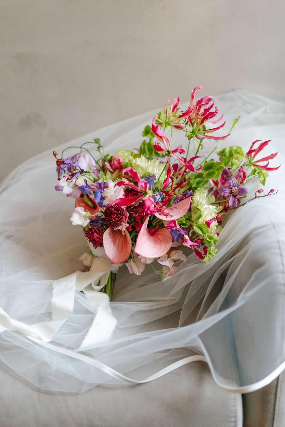 Vibrant bridal bouquet with red gloriosa lilies, pink anthuriums, and purple flowers resting on white tulle fabric.